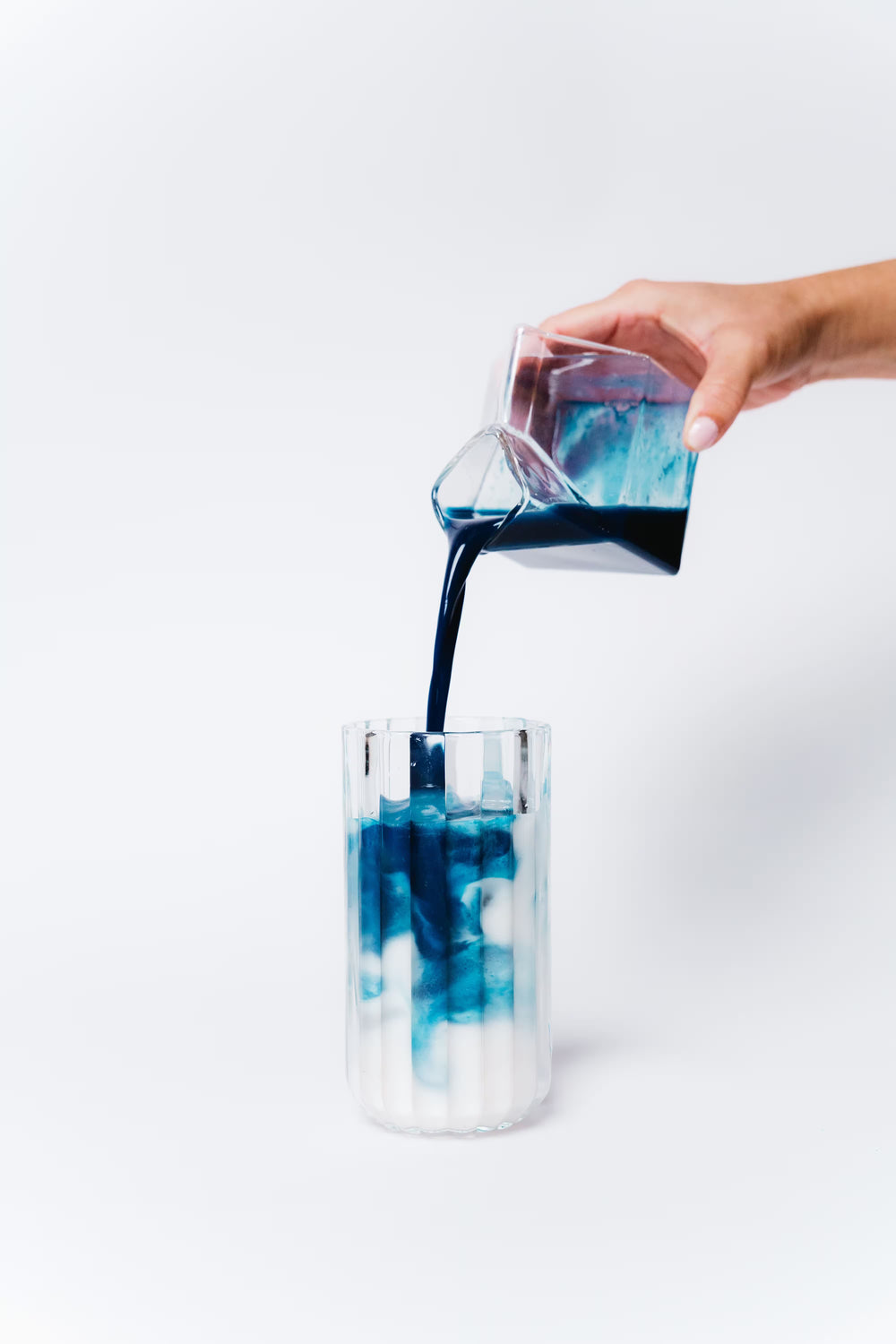 Hand pouring a dark liquid into a glass with blue and white ice cubes on a white background