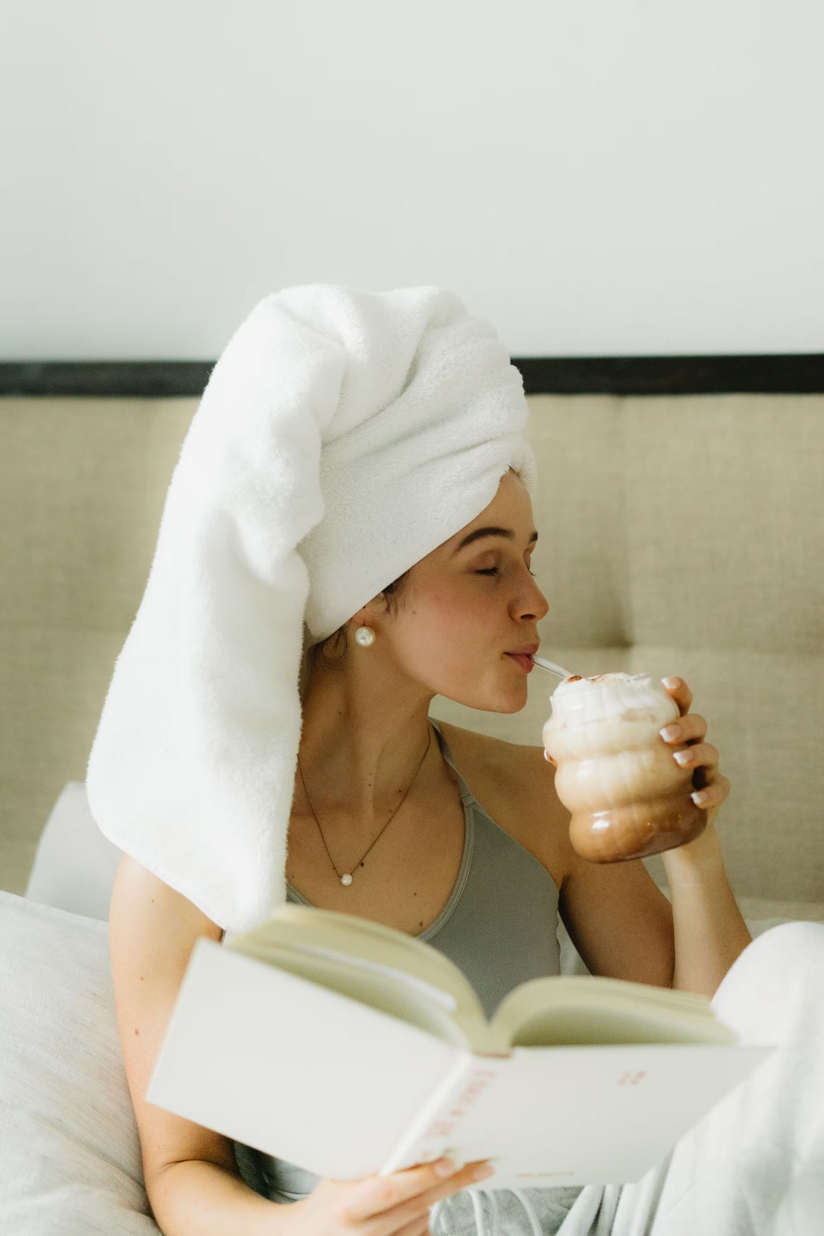 Woman with a towel on her head reading a book and drinking from a cup.