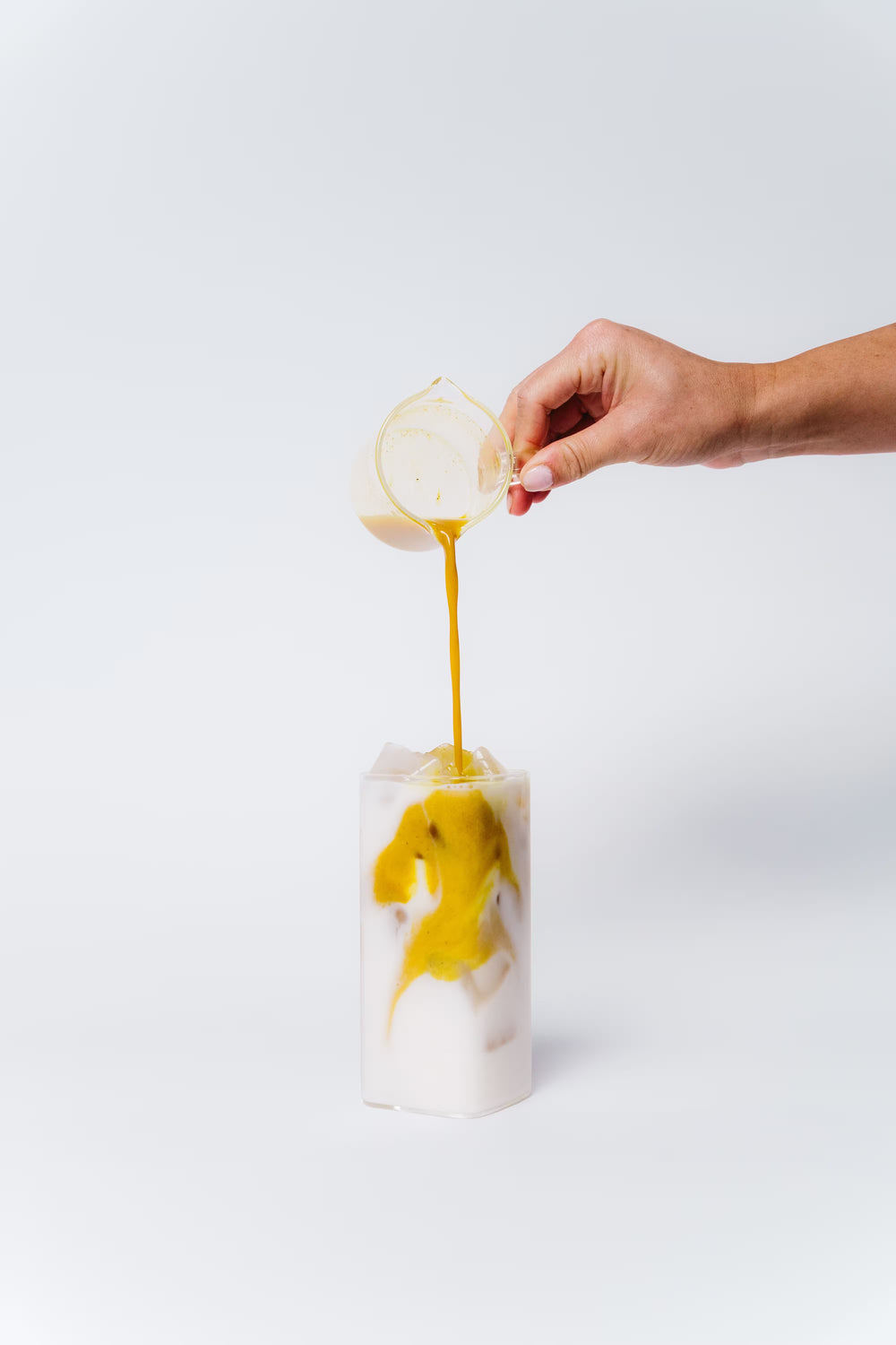 Hand pouring a yellow liquid into a glass with white substance on a white background