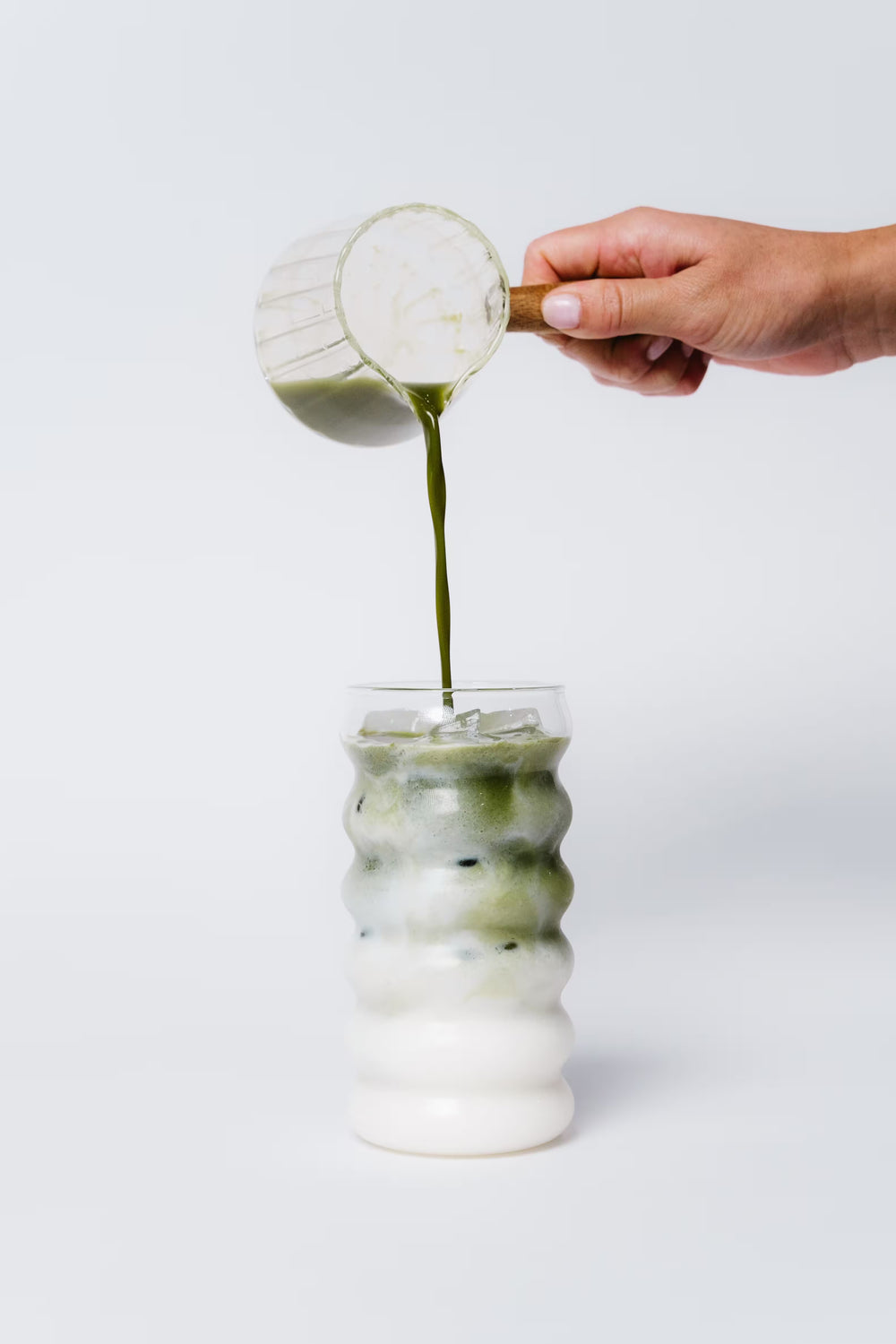 Hand pouring a green liquid from a small container into a glass with a white background
