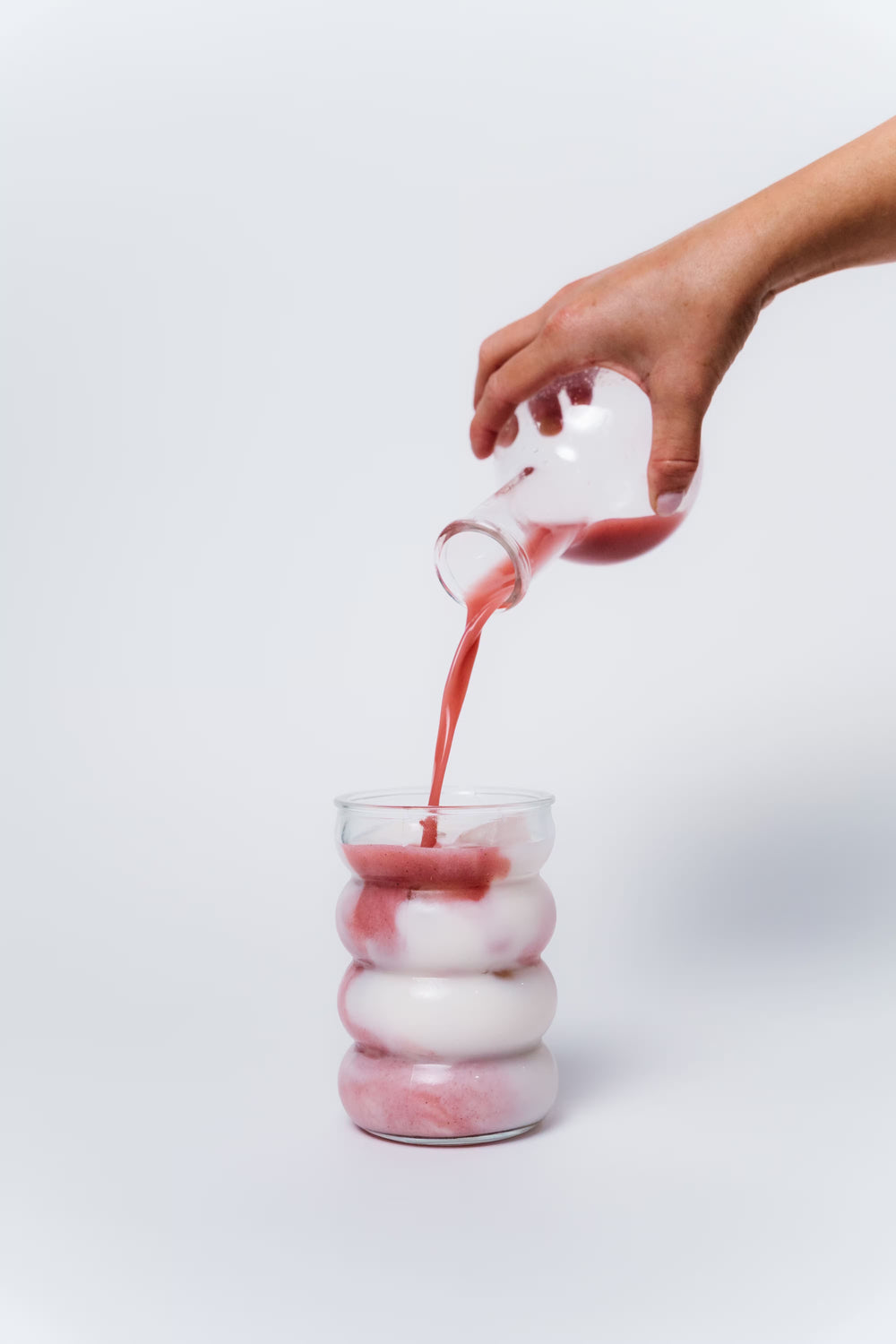 Hand pouring a pink liquid into a glass with stacked white and pink layers on a light gray background
